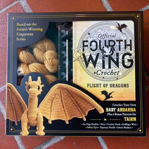 Boxed crochet kit titled 'Official Fourth Wing Crochet: Flight of Dragons' shown on a red brick surface. The package includes a crocheted dragon figure, yarn skeins in a clear window, and printed cover art with text about making a baby Andarna.