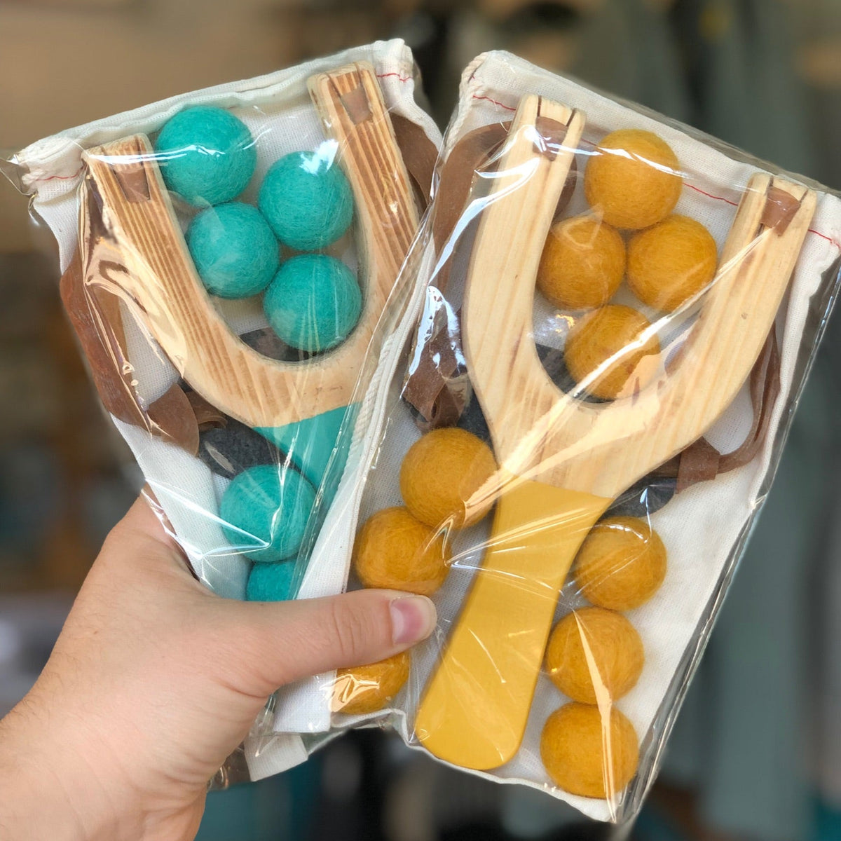 Two packaged wooden slingshots with colorful felt balls, held in a hand against a blurred indoor background.
