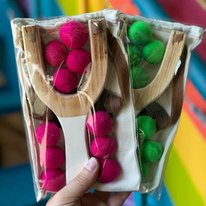 A hand holds a clear plastic bag containing two wooden slingshots with felt balls in bright pink and green, against a colorful blurred background.
