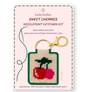 A needlepoint keychain kit featuring a design of two cherries on a white background with a green border. 