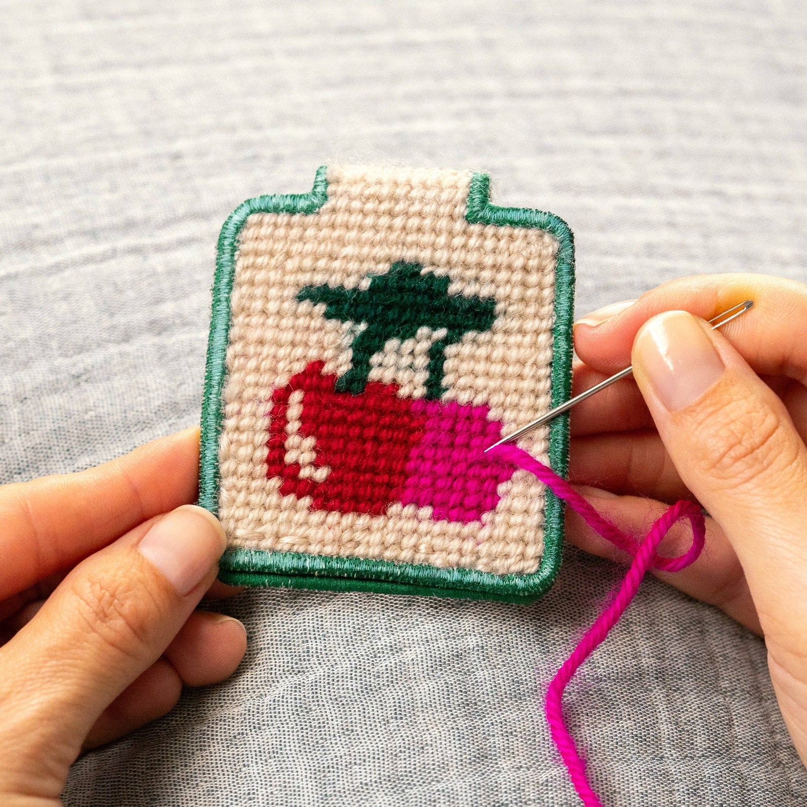 Hand holding a small needlepoint keychain with a red cherry design on a gray fabric background.
