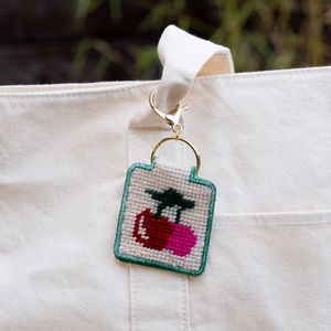 Needlepoint keychain with a cherries design attached to a white canvas bag. 