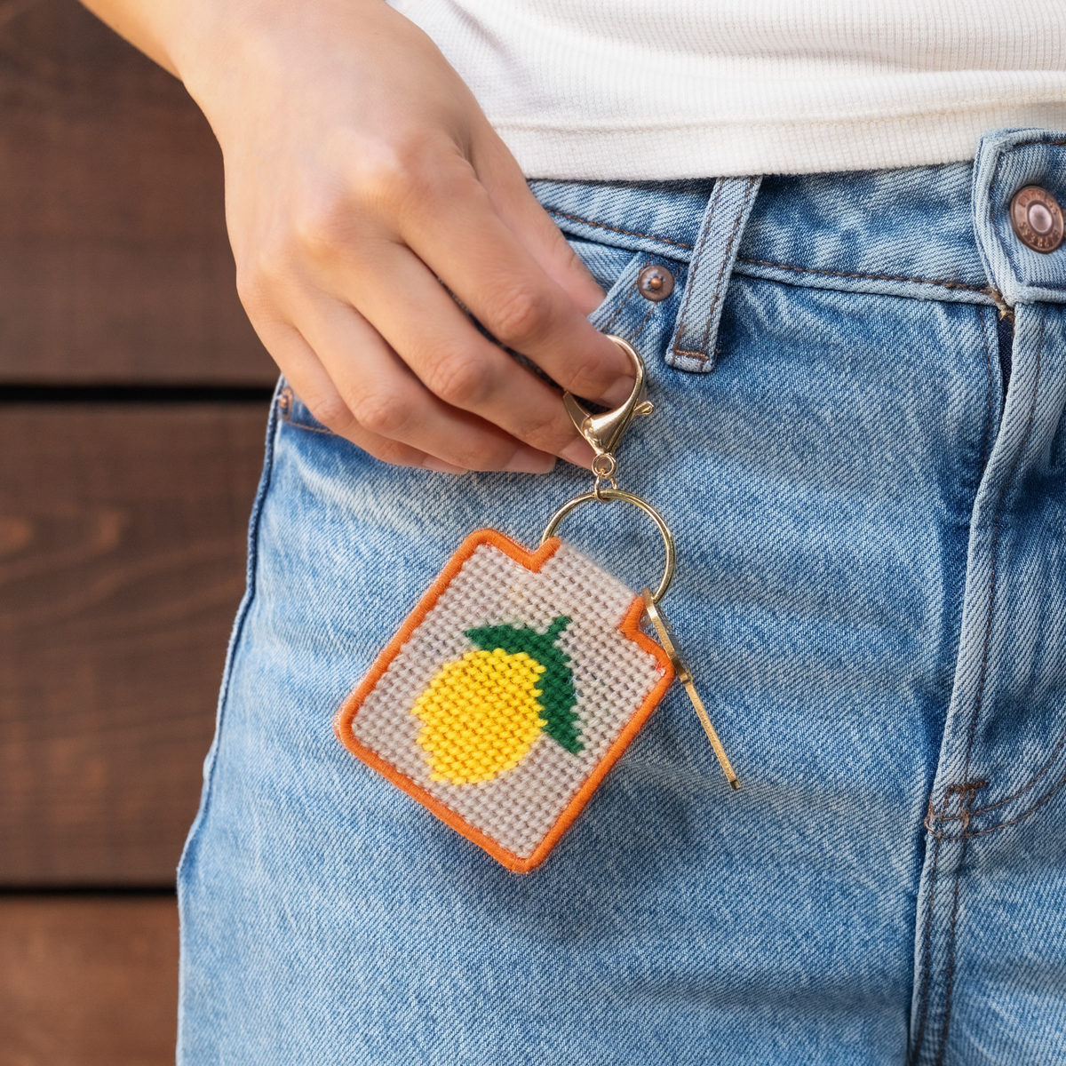 A person wearing jeans holding a lemon needlepoint keychain with a key attached. 