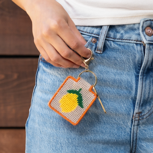 A person wearing jeans holding a lemon needlepoint keychain with a key attached. 