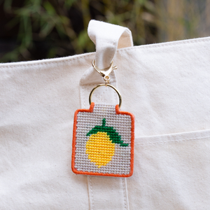 Needlepoint keychain with a lemon design attached to a white canvas bag. 