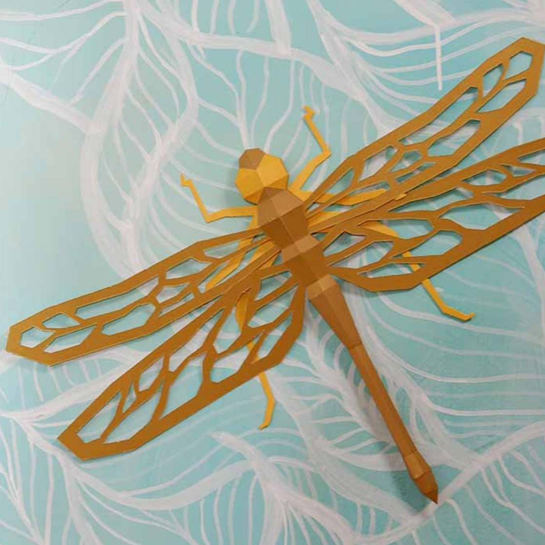 Example of a finished dragonfly paper craft. It is kraft colored paper and it is sitting on top of a mint colored background with white leaves painted on it.
