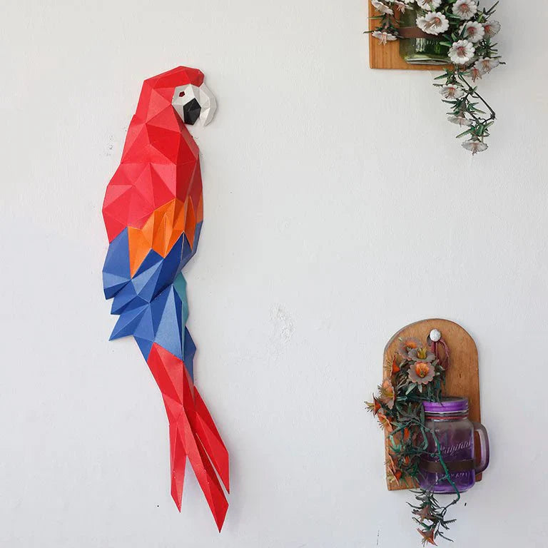 A finished example of a macaw paper craft kit. It is hanging on a wall next to some flowers in mason jars.