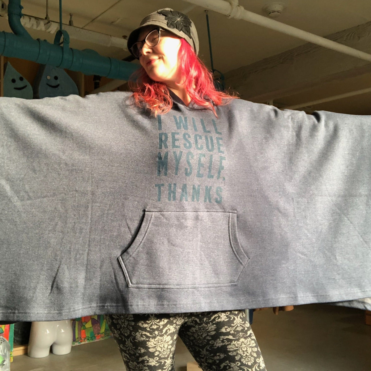 Person wearing a large navy blue hooded poncho holds out her arms to show how wide the poncho is.  The text on the poncho says, "I will rescue myself, thanks."