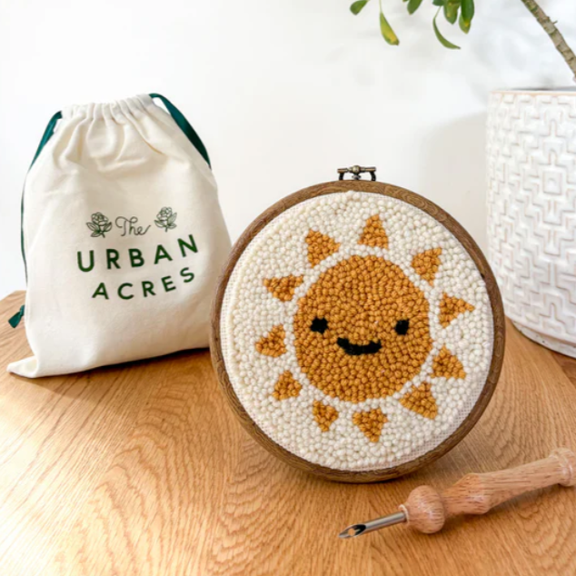 A completed punch needle kit of a happy, smiling sun, is sitting on a table. There is a cloth bag with the vendor name - The Urban Acres - in the background and a wooden punch needle in the foreground.