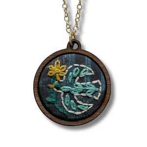 A round wooden pendant necklace featuring an embroidered design of a white dove with teal and yellow accents on a dark textured fabric background, suspended from a gold chain.
