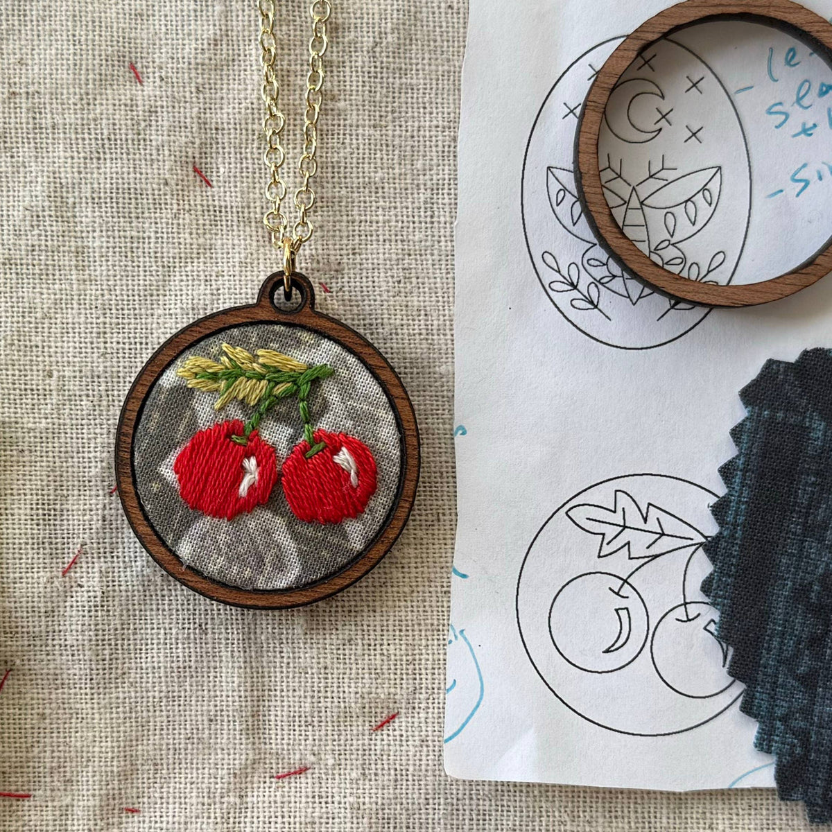 Embroidered pendant with red cherries on a fabric background with embroidery tools.