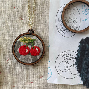 Embroidered pendant with red cherries on a fabric background with embroidery tools.