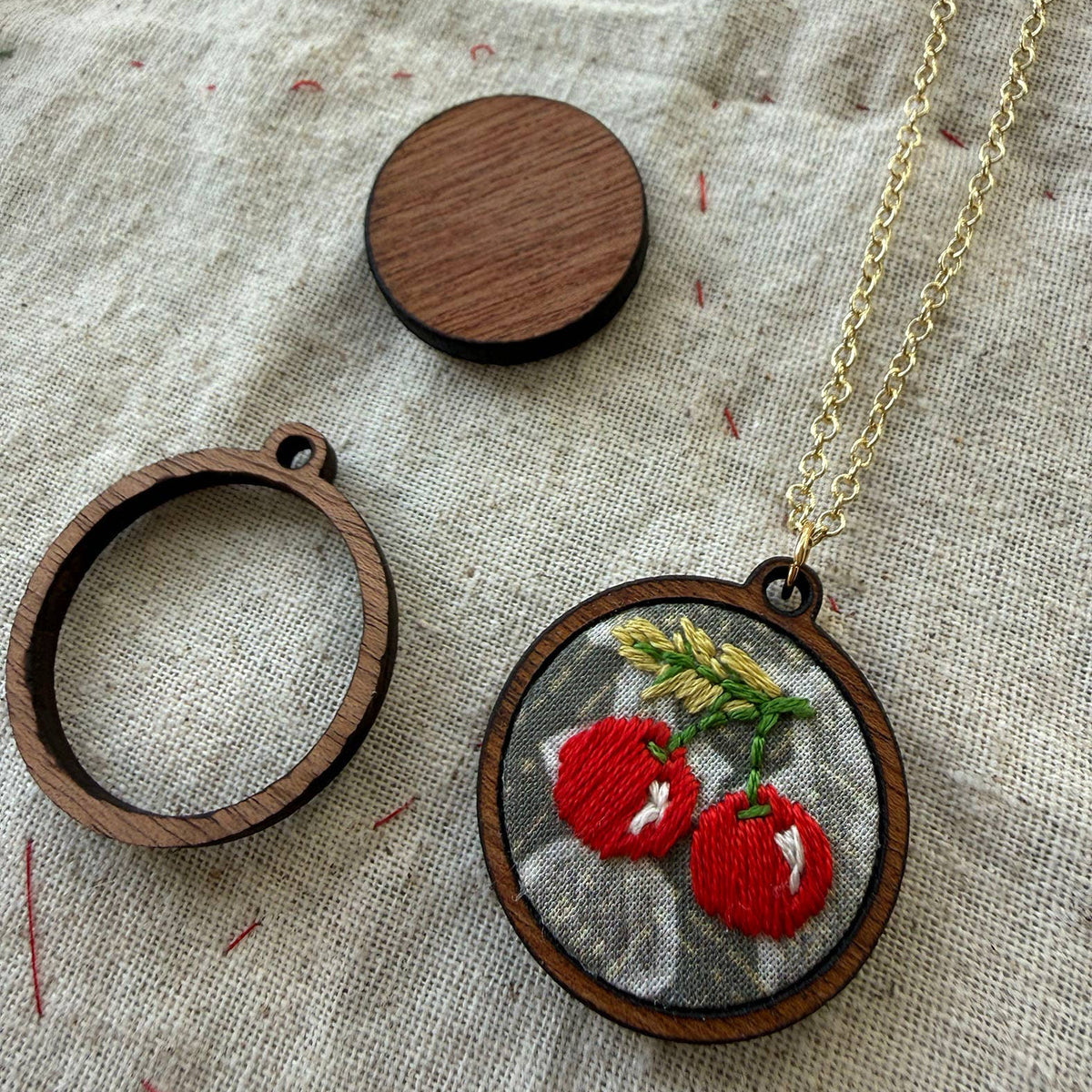 Embroidered pendant with red cherries on a wooden hoop, gold chain, and wooden disc on fabric background
