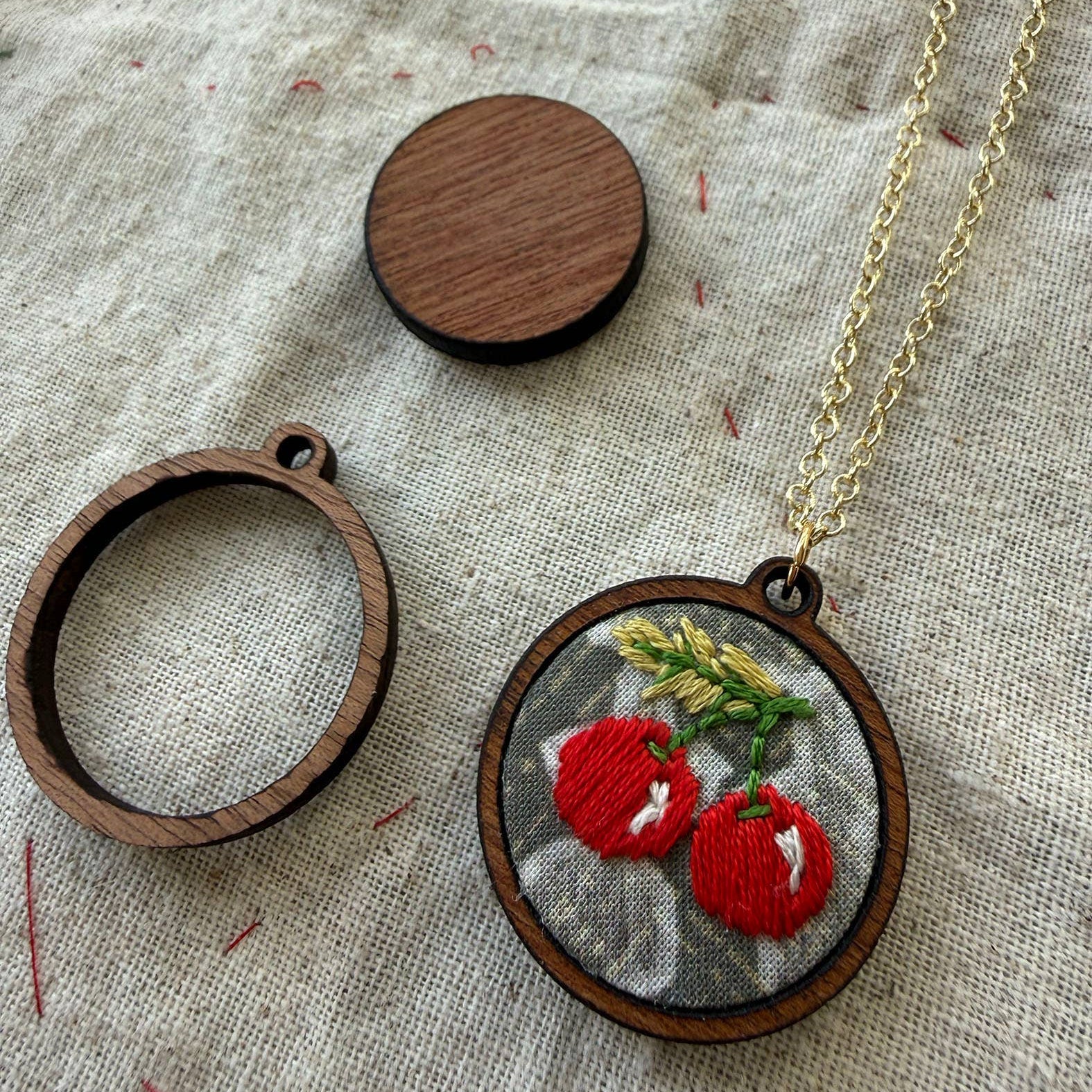 Embroidered pendant with red cherries on a wooden hoop, gold chain, and wooden disc on fabric background