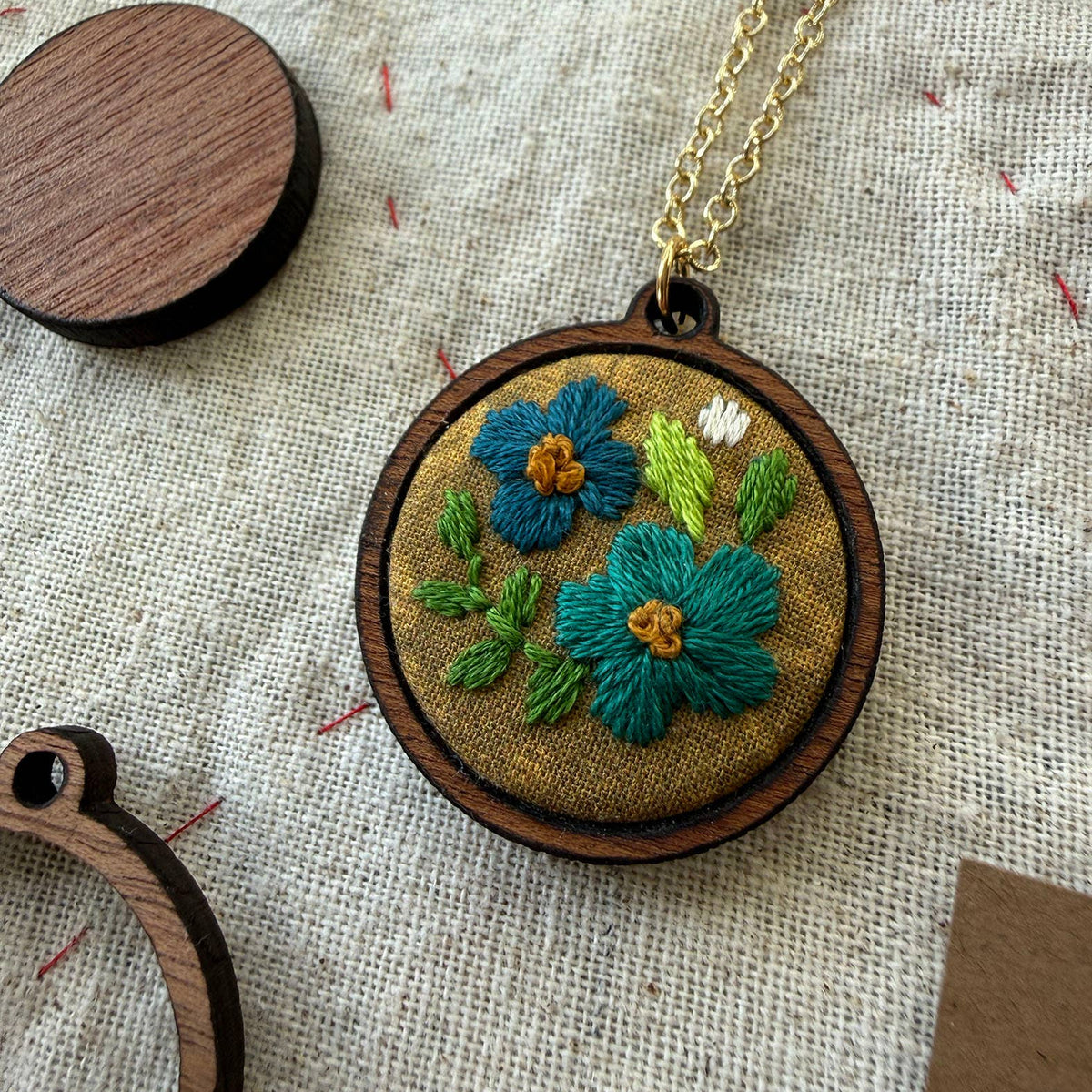 Embroidered necklace with floral design on a wooden hoop, placed on a textured fabric background.