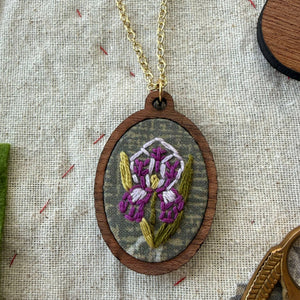 Embroidered pendant with a floral design on a chain, placed on a textured fabric background.