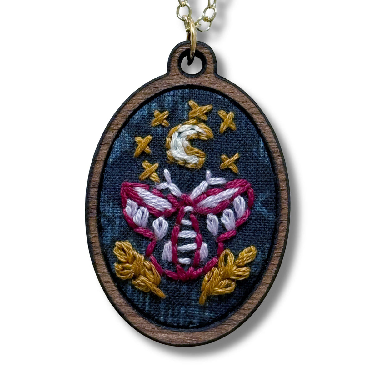 Embroidered pendant with moth design on a wooden frame, hanging on a chain against a white background.