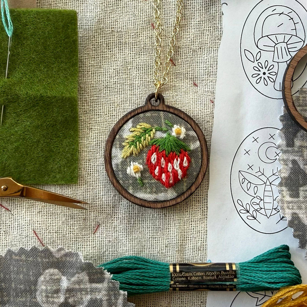 Embroidery hoop with strawberry design on fabric, surrounded by embroidery tools and patterns.