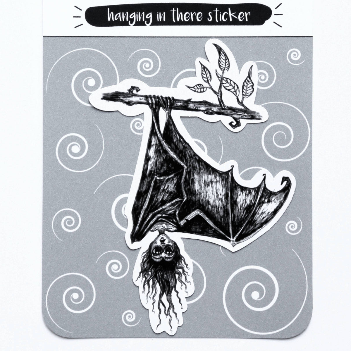 A hanging in there sticker by Scream Print, featuring a black and white illustration of a bat hanging from a branch with a woman's face, presented on its gray backing card.