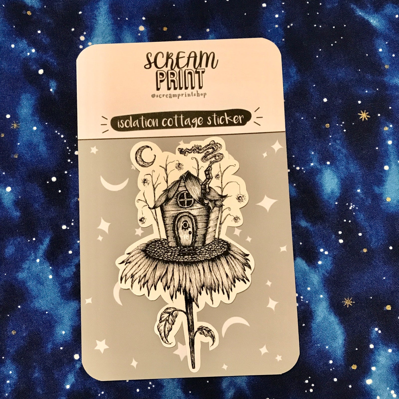 A sticker on a branded backing card of a tiny cottage on top of a sunflower with smoke coming out of the chimney and a crescent moon in the background. 