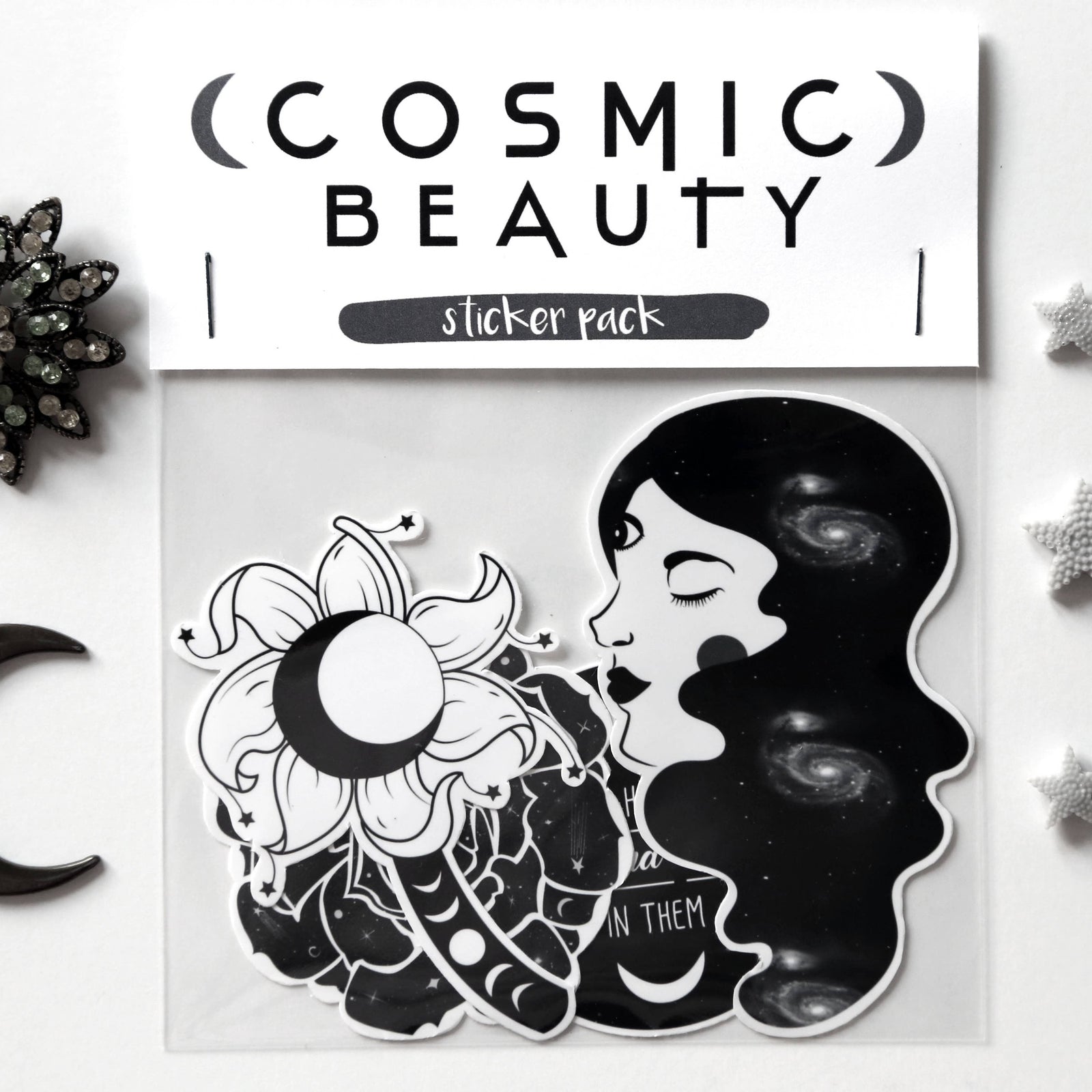 A sticker pack labeled 'COSMIC BEAUTY sticker pack' shown on a white background with decorative celestial props including a black crescent moon, a rhinestone flower brooch, and silver star embellishments. The sticker set includes black-and-white illustrated stickers featuring a woman's profile, a flower, and moon motifs.