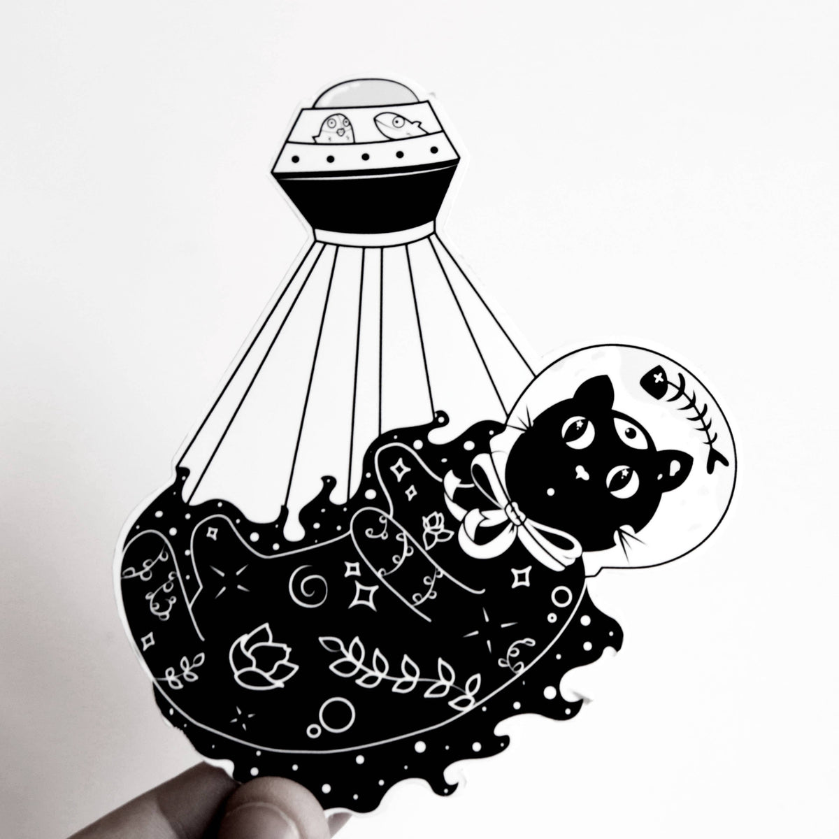 A black and white sticker featuring a cute cartoon cat in a space suit floating under a UFO, held by a hand against a plain white background.