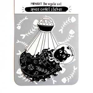 A product card for a 'Scream Print' sticker titled 'Midnight the Mystic Cat space cadet sticker'. The card features a black and white illustration of a cat beneath a UFO, surrounded by fish bones on a light gray background.