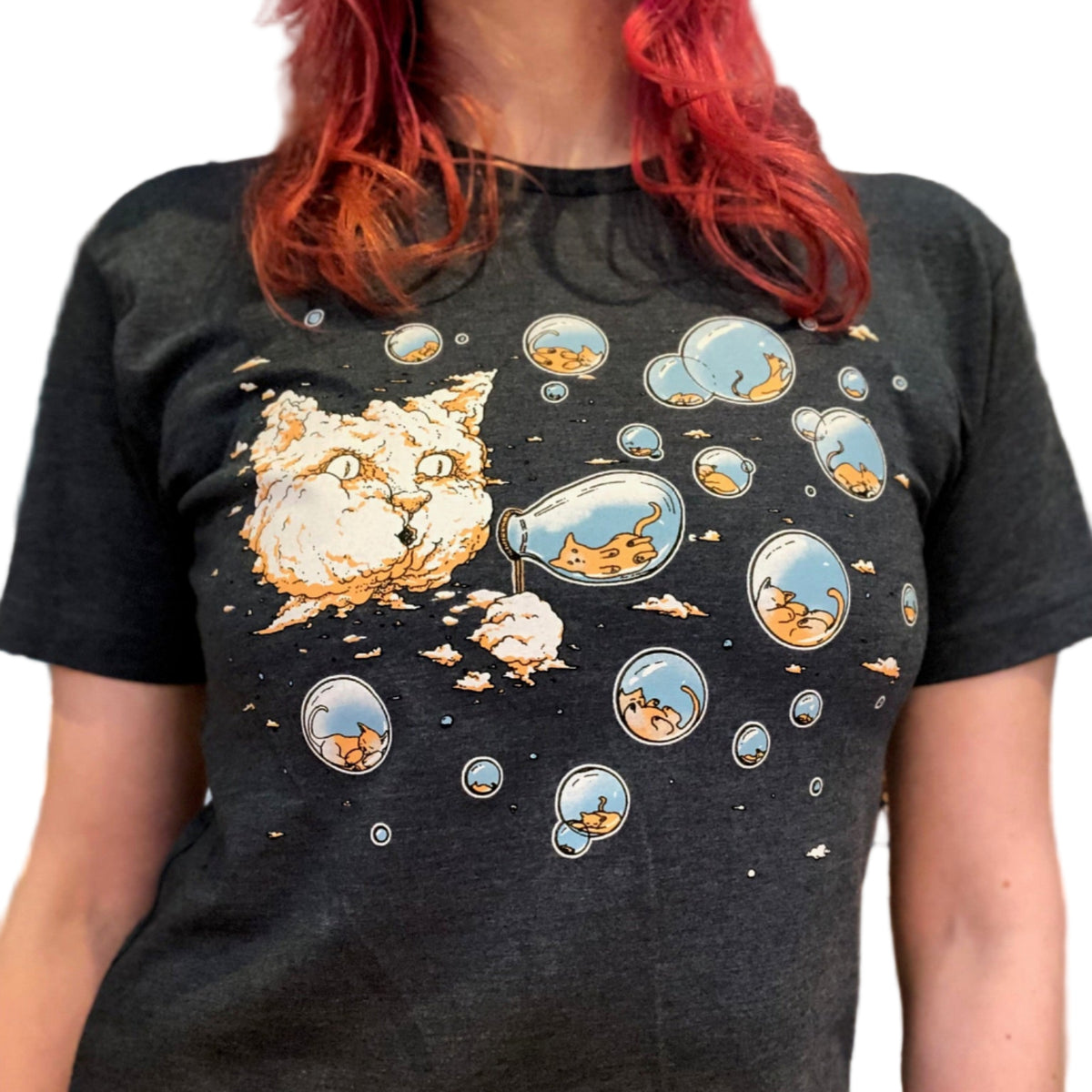 Person wearing a blue t-shirt with cartoon cat designs. There is a large cloud in the sky that looks like a giant cat head. The cat head is blowing bubbles that have tiny kitties inside. 