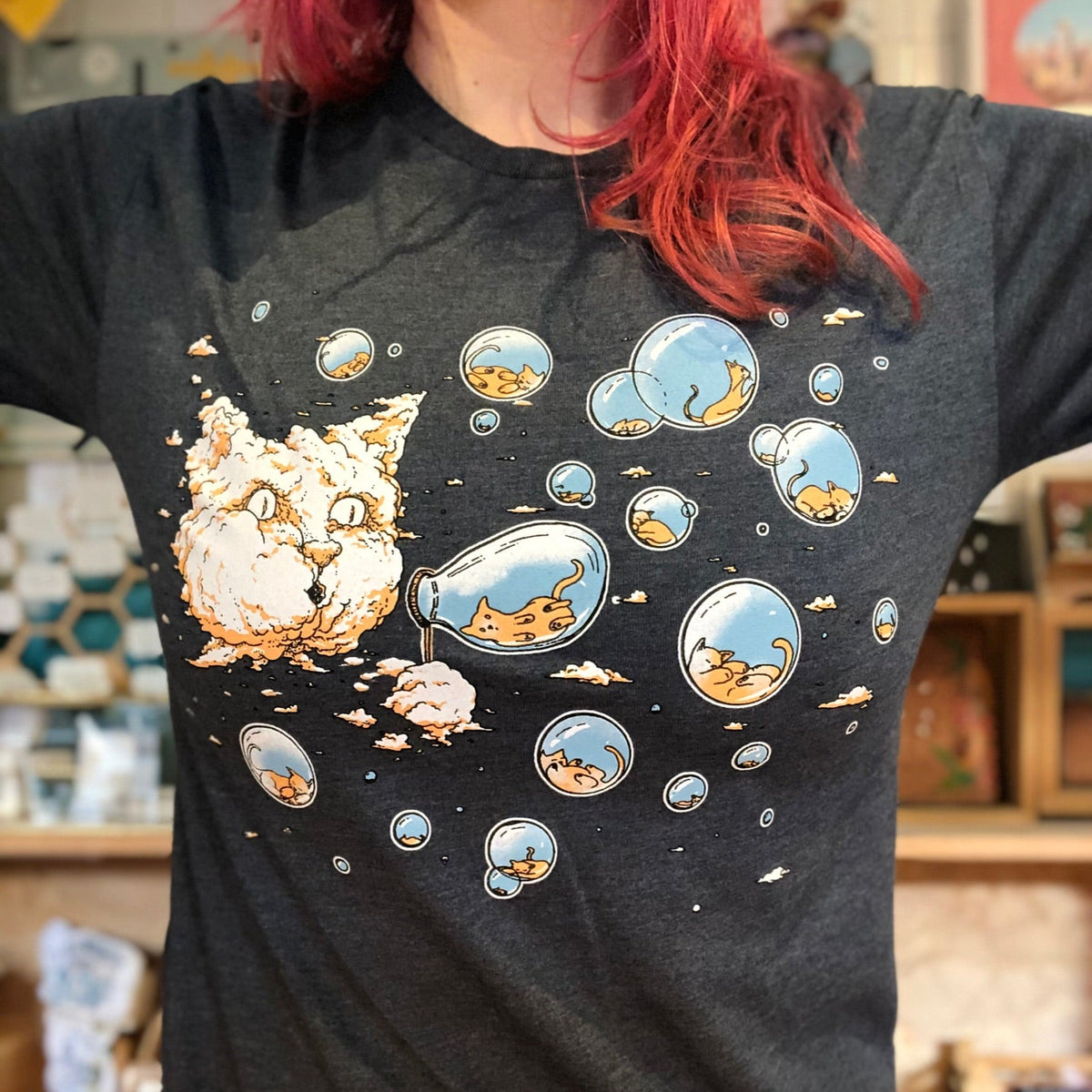 Person wearing a blue t-shirt with cartoon cat designs. There is a large cloud in the sky that looks like a giant cat head. The cat head is blowing bubbles that have tiny kitties inside. 