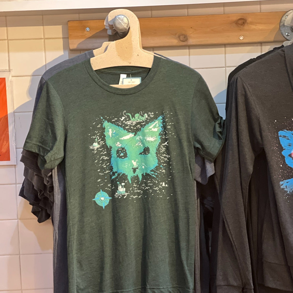 A green catographer design tshirt in a store setting hanging on a wood rack.