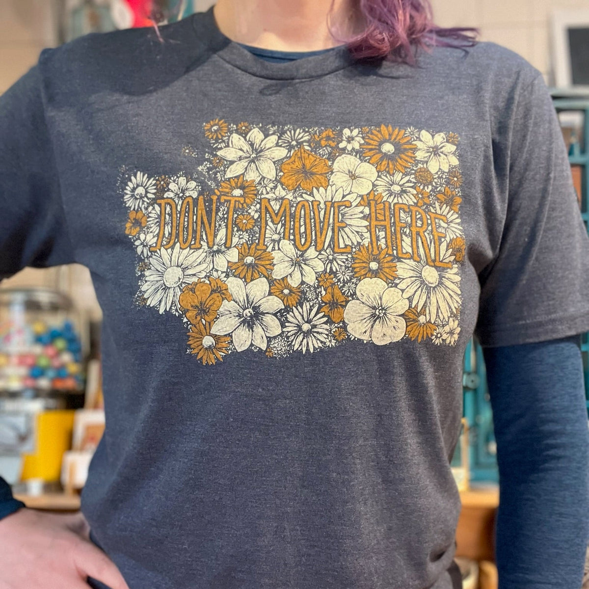 Person wearing a blue t-shirt with floral design and text that says "Don't Move Here" in a store setting. The floral design is in the shape of Washington state. 
