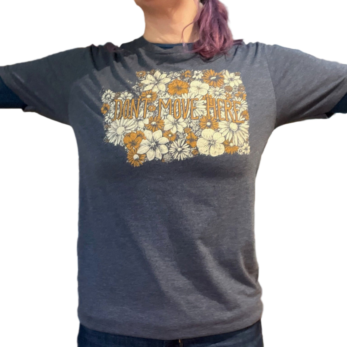Person wearing a blue t-shirt with floral design and text that says "Don't Move Here.". The floral design is in the shape of Washington state. 