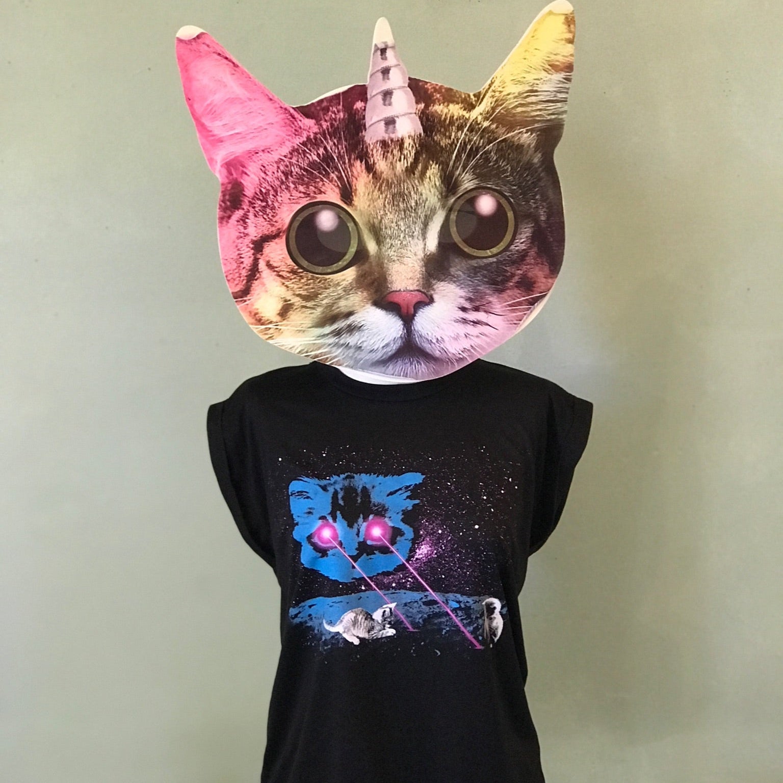 A mannequin wearing a unicorn-cat-head mask is displaying a rolled cuff muscle tee with our Meta Laser Cat design. A blue cat head is floating in space and shooting hot pink lasers out of his eyes. Kittens are playing in the lasers. 