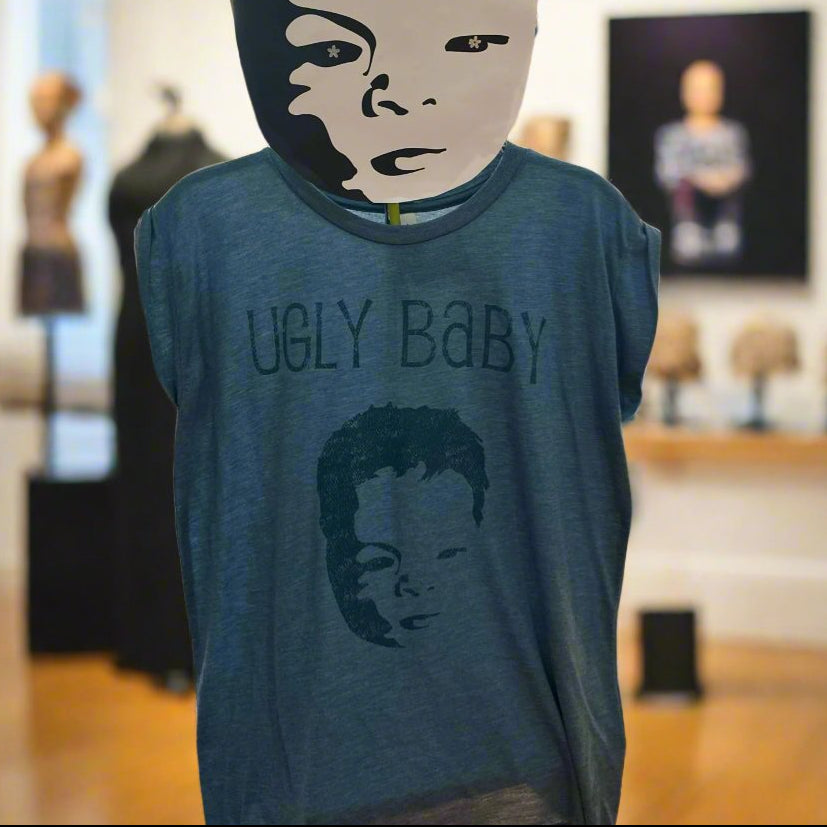 A cap sleeve shirt displayed on a mannequin in a museum setting, featuring the text 'UGLY BABY' with a stylized baby face design.