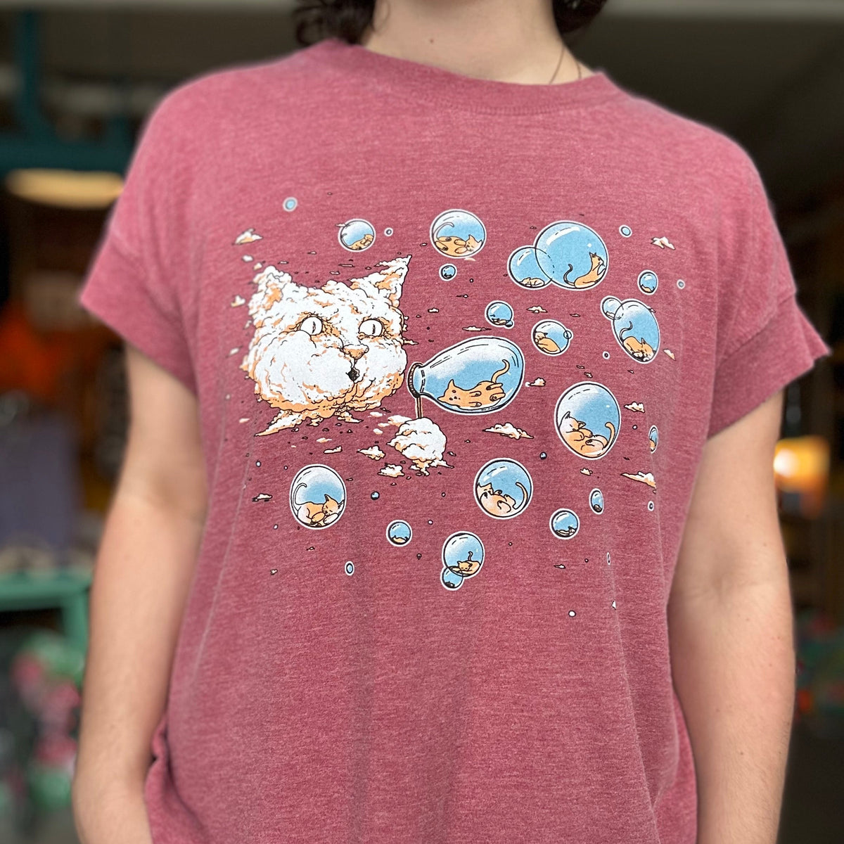 Person wearing a maroon t-shirt with a cartoon cat design. There is a cloud in the shape of a large cat head that is blowing bubbles. Inside the bubbles are tiny kittens.