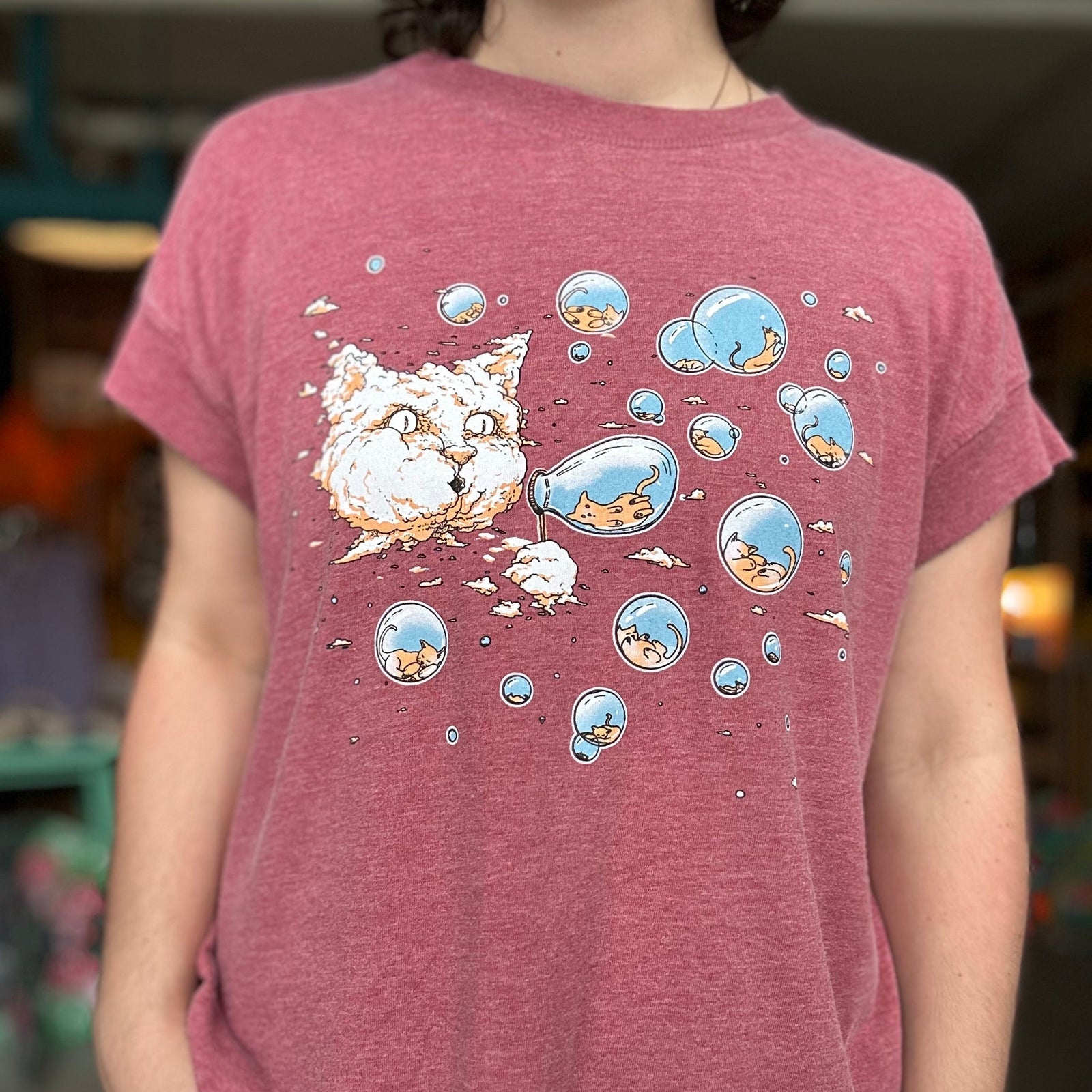 Person wearing a maroon t-shirt with a cartoon cat design. There is a cloud in the shape of a large cat head that is blowing bubbles. Inside the bubbles are tiny kittens.