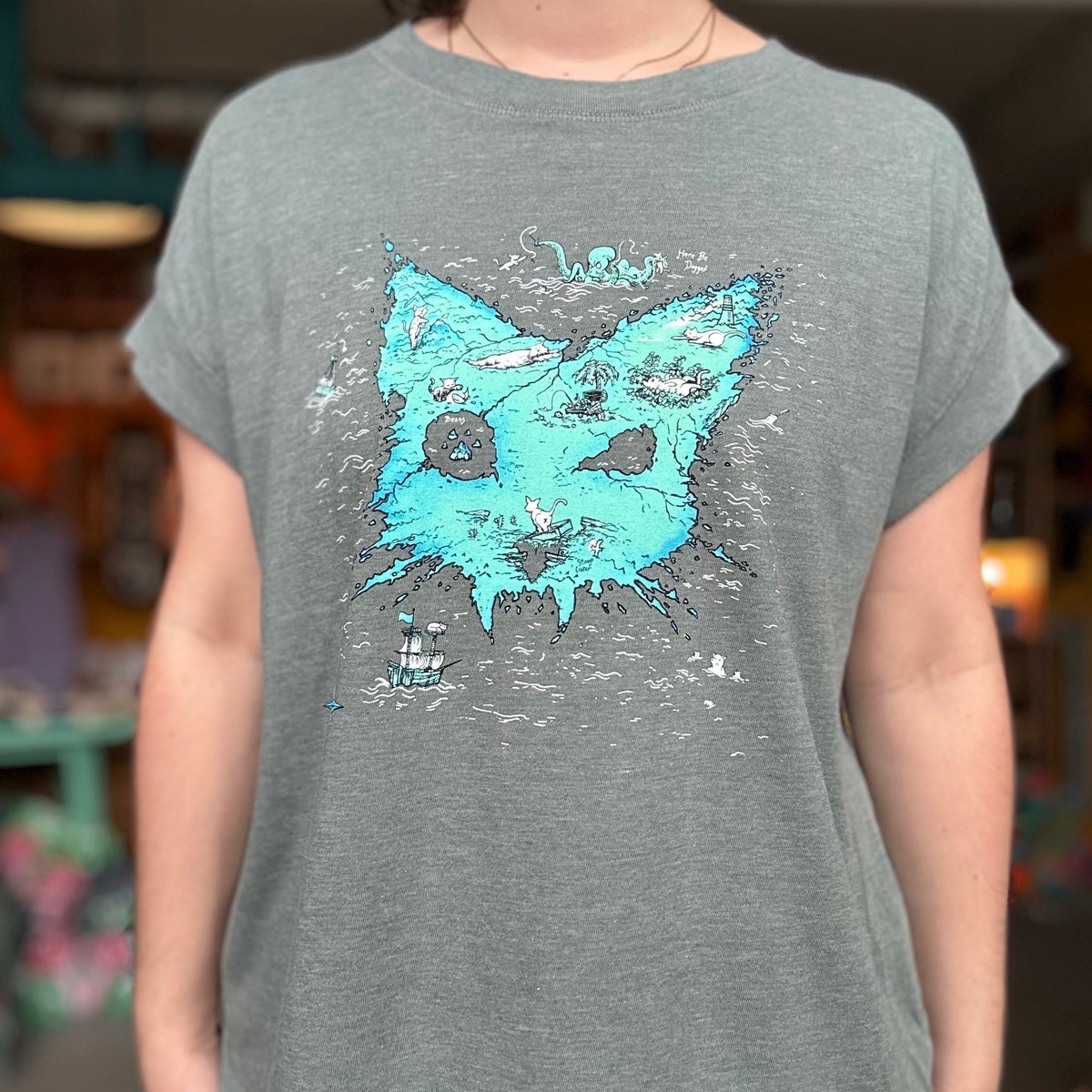 Person wearing a heather basil t-shirt with the Catographer design. A treasure map of an island that looks like a cat head. 