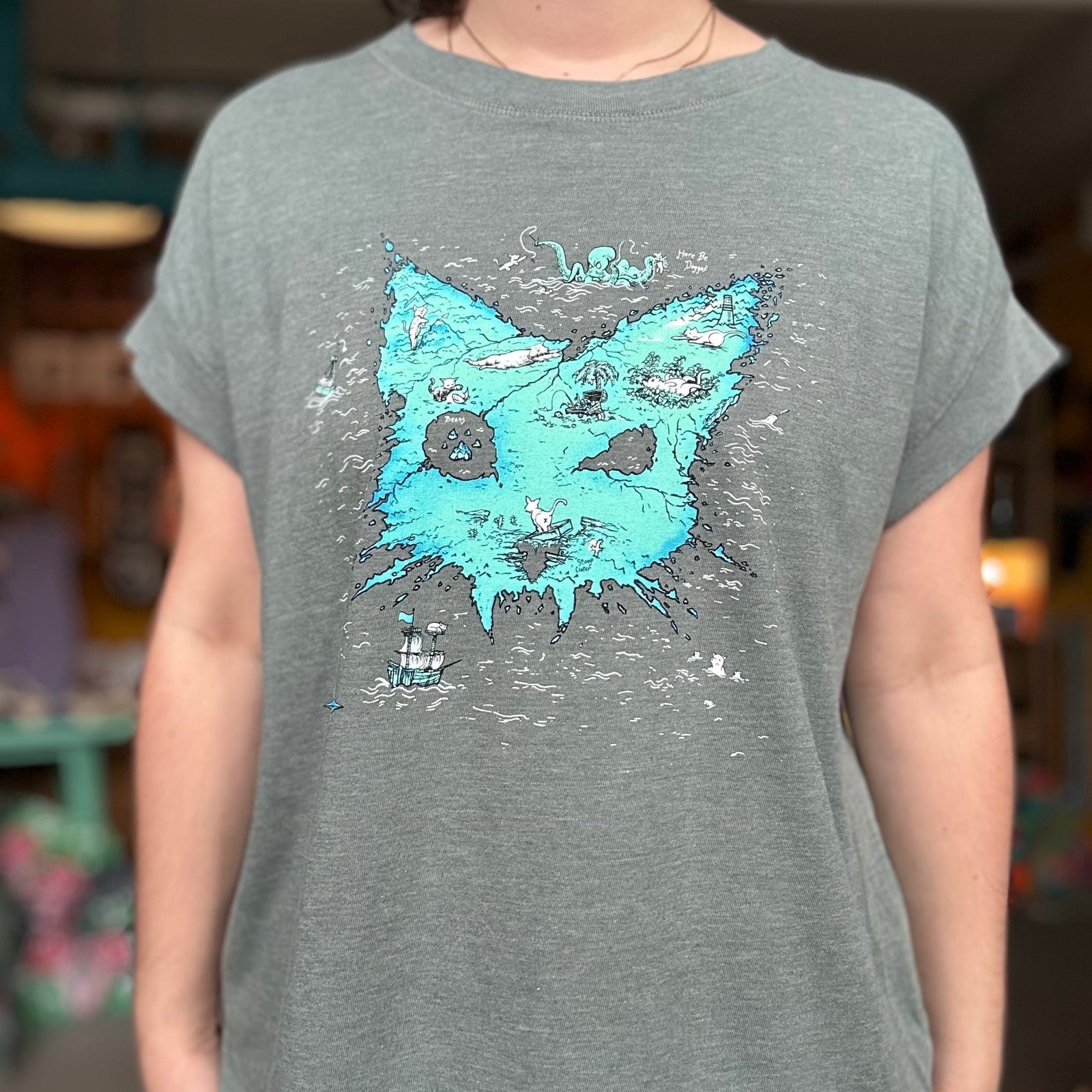 Person wearing a heather basil t-shirt with the Catographer design. A treasure map of an island that looks like a cat head. 