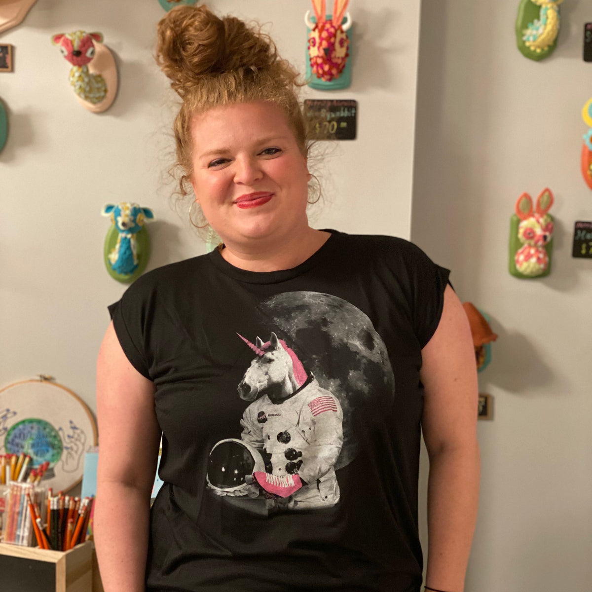 A person wearing a black flowy rolled cuff muscle tee with an astronaut unicorn screenprinted on it in white and hot pink. 