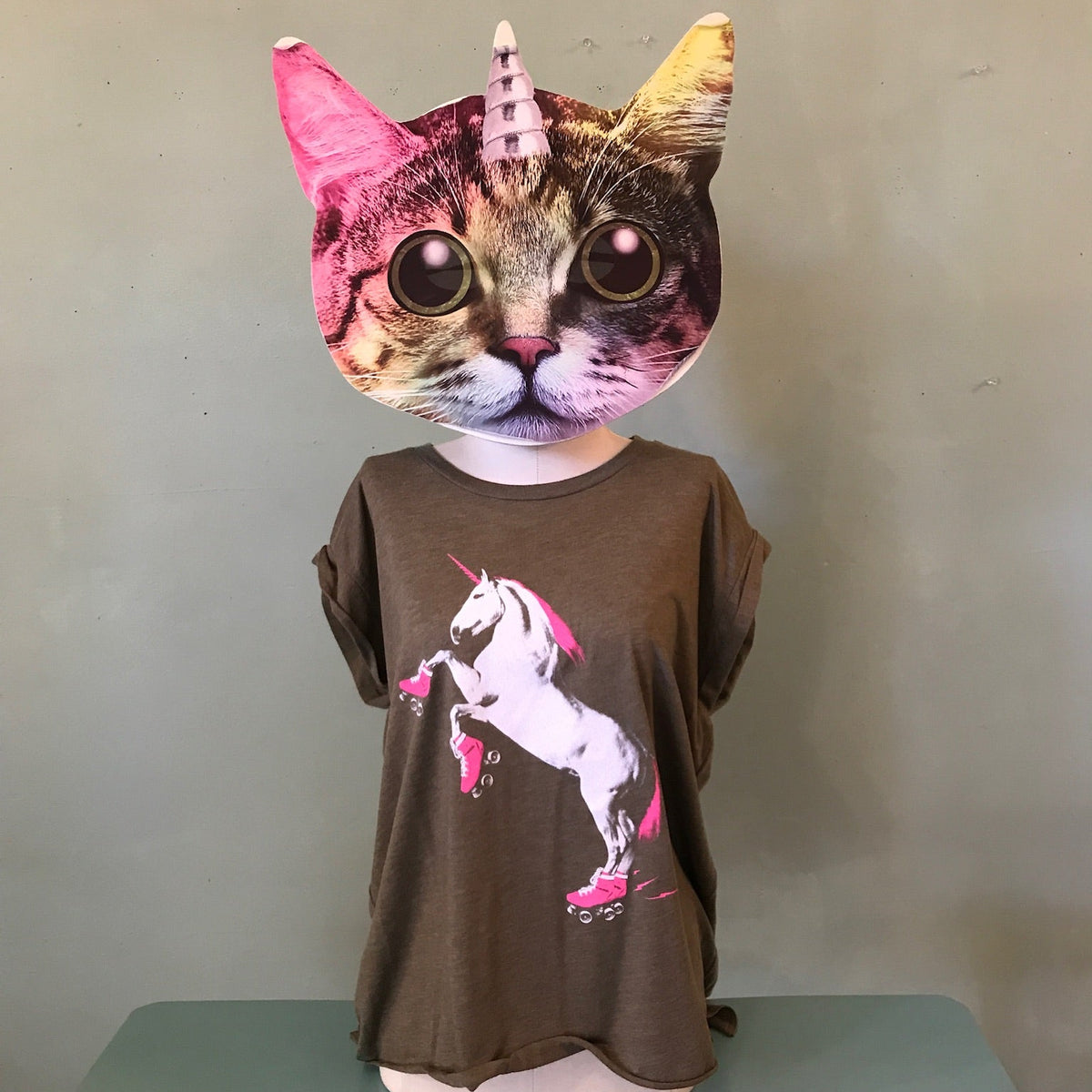 Person wearing a cat mask with a colorful unicorn horn and a brown shirt with a roller skating unicorn design in neon pink and white. 