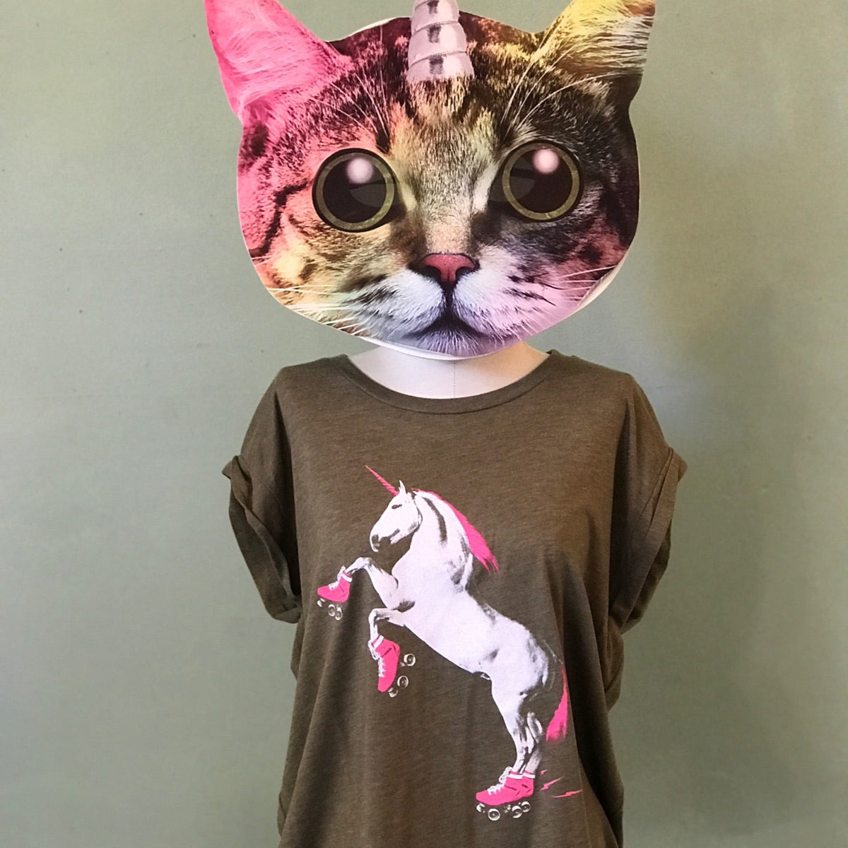 A cat-unicorn mask on a feminine mannequin form is wearing a rolled cuff muscle tee in heather olive with a neon pink and white roller skating unicorn on the front. 