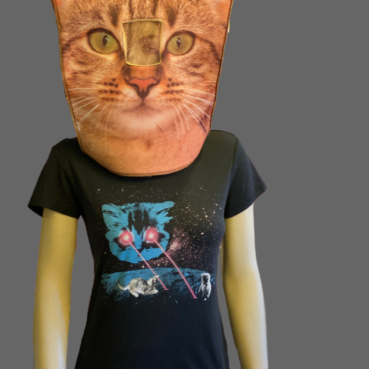Mannequin (with the head of a giant cat) is wearing a black t-shirt with a meta laser cat graphic. There is a large blue cat head in the sky shooting hot pink lasers onto the surface of the moon. Tiny kittens are playing in the lasers. 