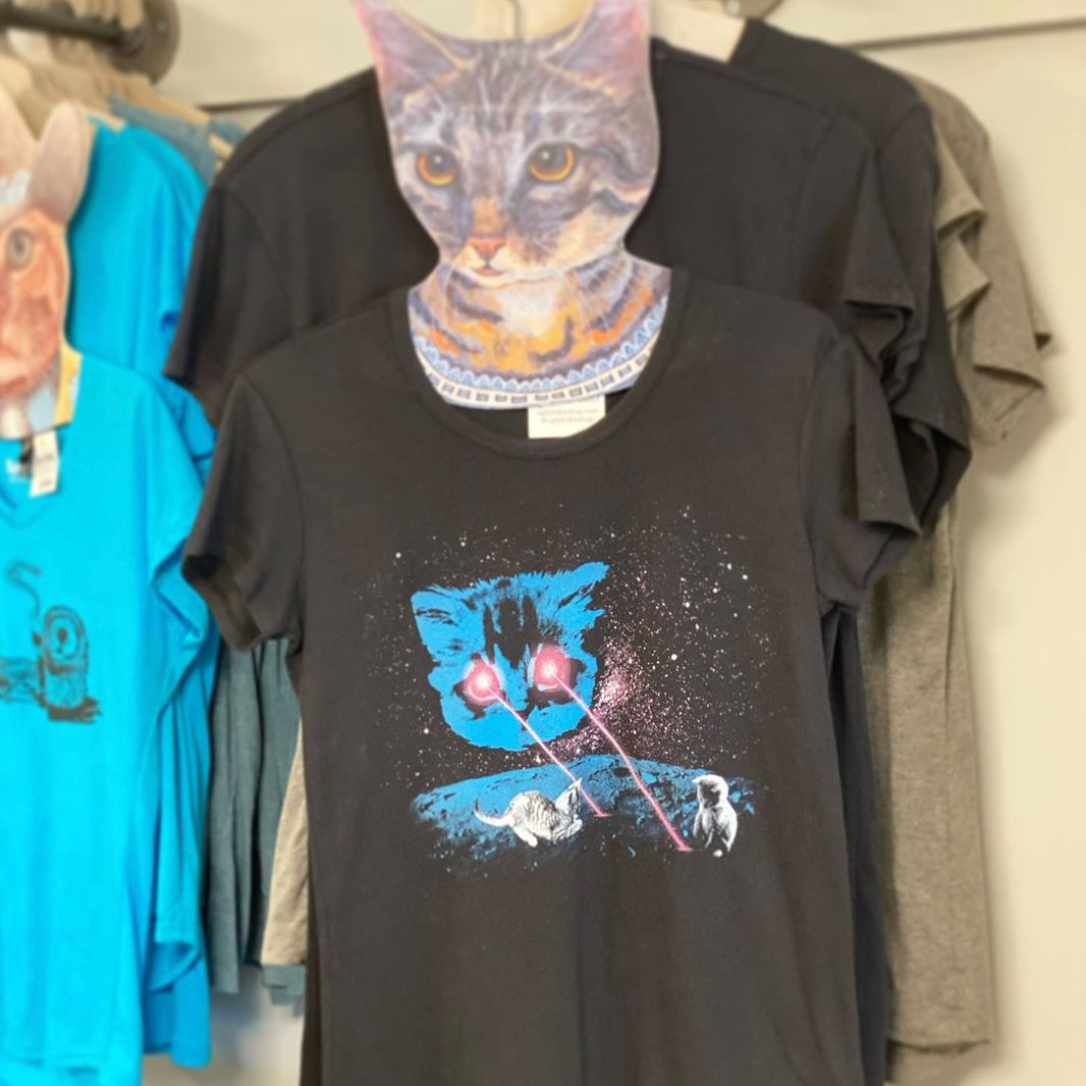 Black t-shirt with a cat-themed graphic design hanging on a rack with other shirts.