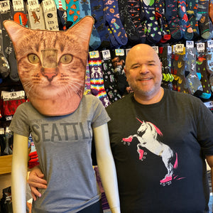 Man standing next to a mannequin wearing a cat mask in a store with various products on shelves. The mannequin is wearing a shirt that says "This Says Seattle On It." The man is wearing a Roller Skating Unicorn shirt. 