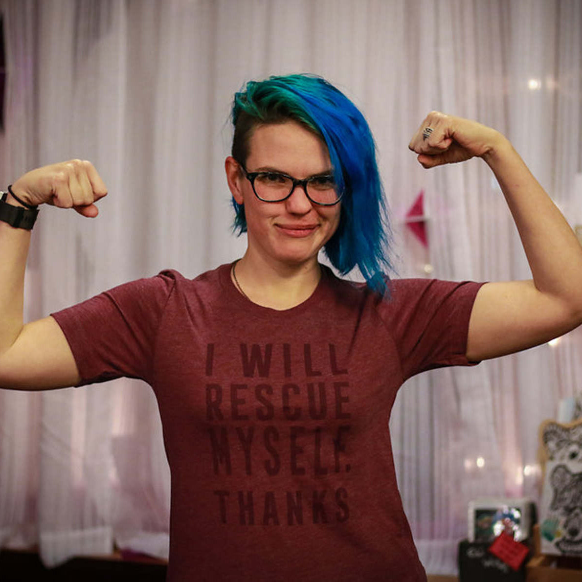 Person with blue hair wearing a maroon t-shirt with text that says "I will rescue myself, thanks", flexing muscles indoors.