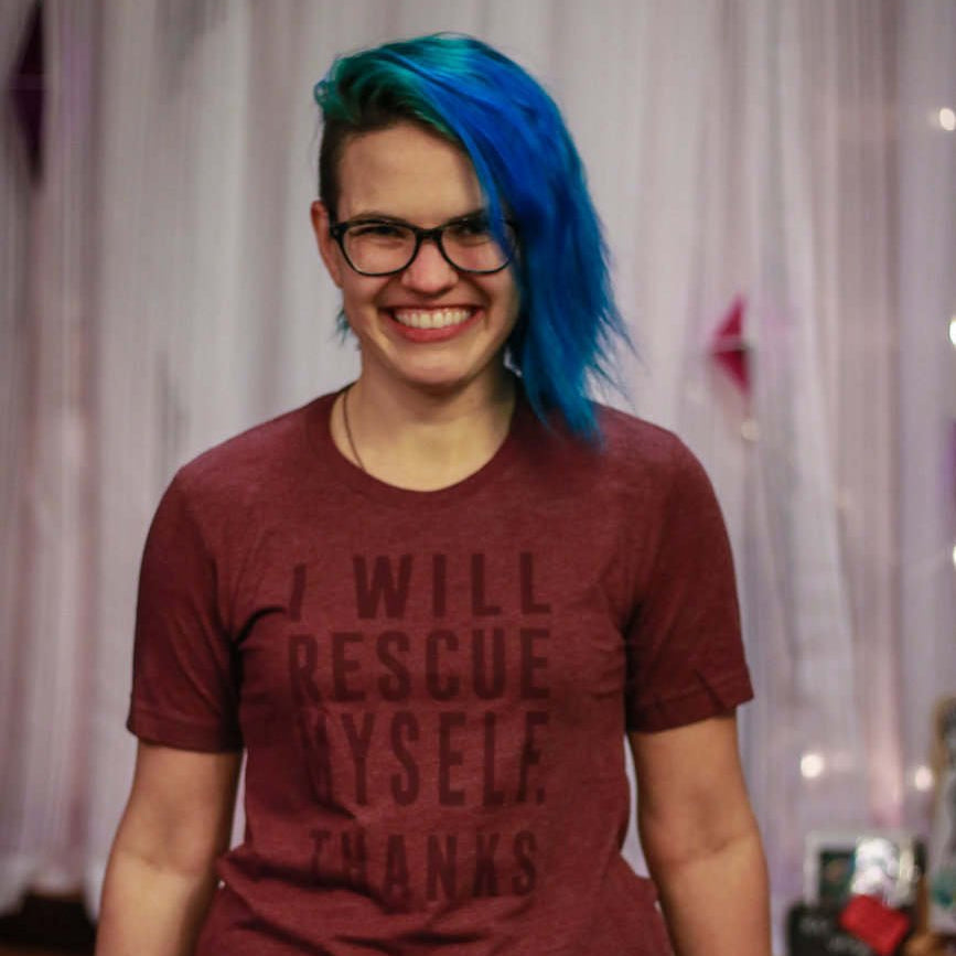 Person with blue hair wearing a maroon t-shirt with text  that says "I will rescue myself, thanks", smiling in front of a curtain.