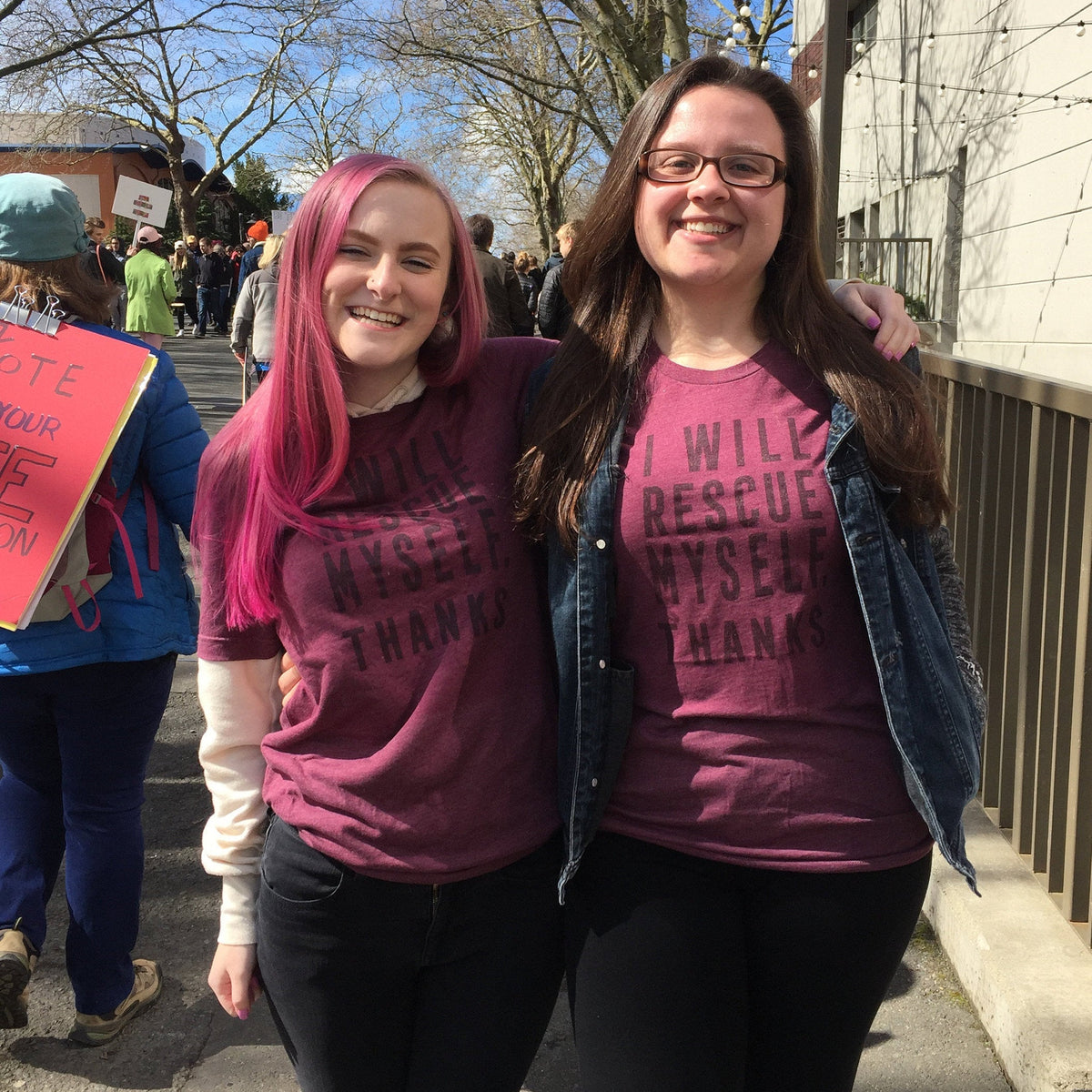Two women wearing matching maroon t-shirts with text that says "I will rescue myself, thanks", standing outdoors on a sunny day.