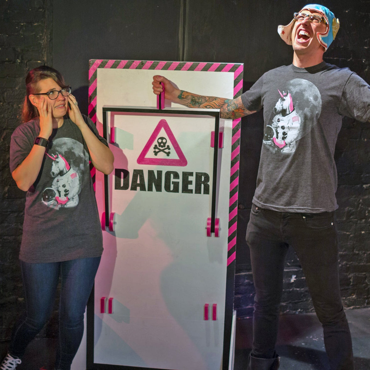 Two people in front of a 'DANGER' sign with a skull and crossbones symbol. They are both wearing astronaut unicorn shirts.