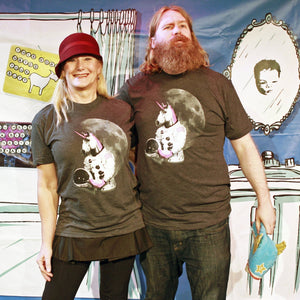 Two people wearing matching t-shirts with a graphic design of an astronaut unicorn, standing in front of a colorful background.