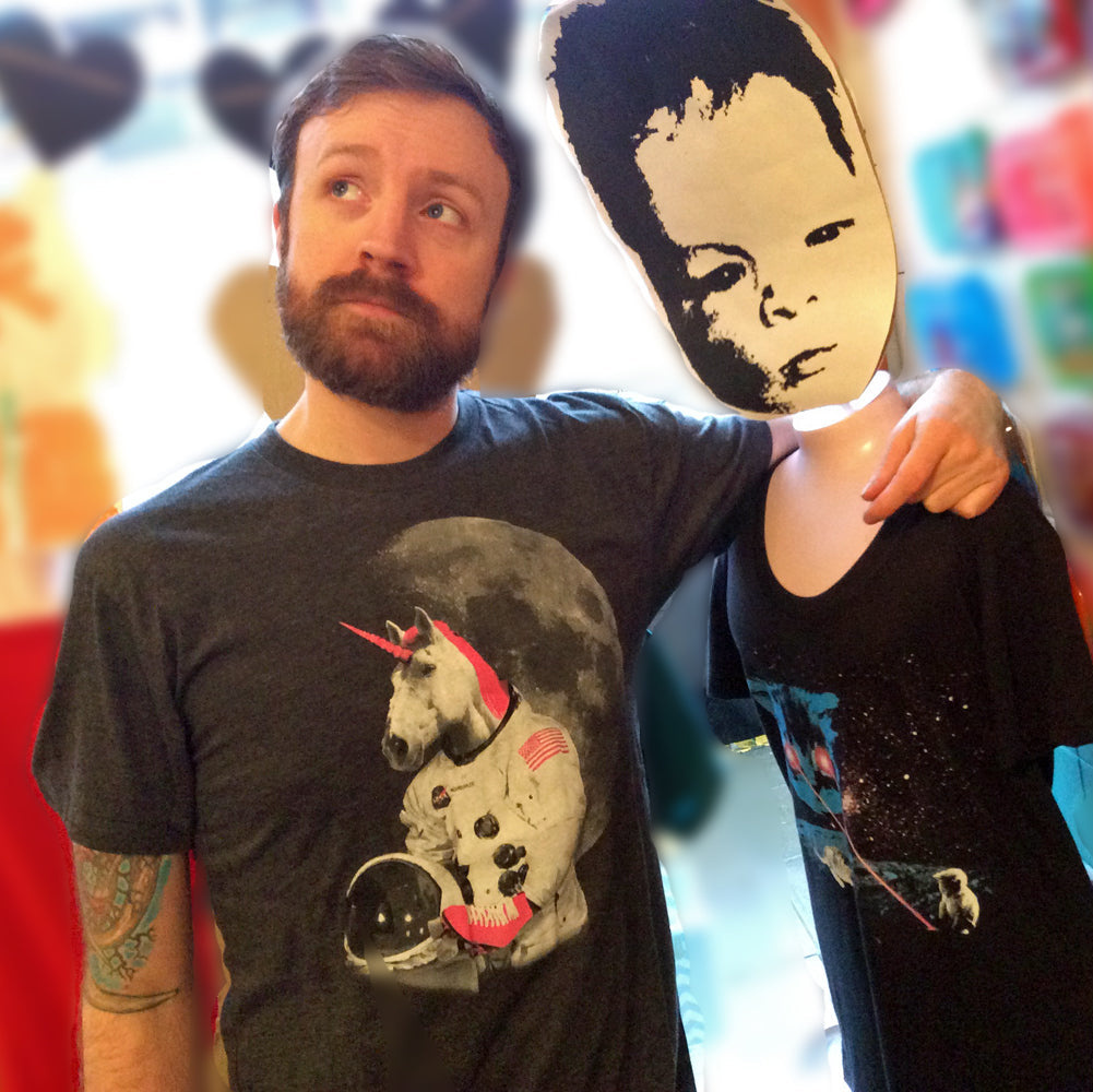 Man with his arm around a mannequin wearing a mask. He is wearing a dark gray tshirt with an astronaut unicorn screenprinted in hot pink and white. 